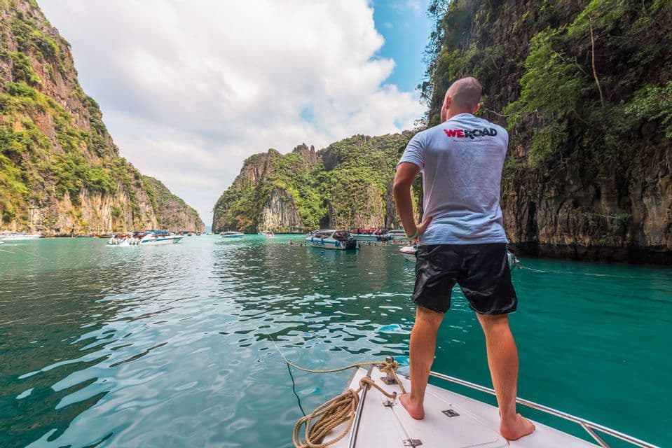 A man from a WeRoad trip stands on the bow of a boat, looking out over a turquoise bay with rocky cliffs.