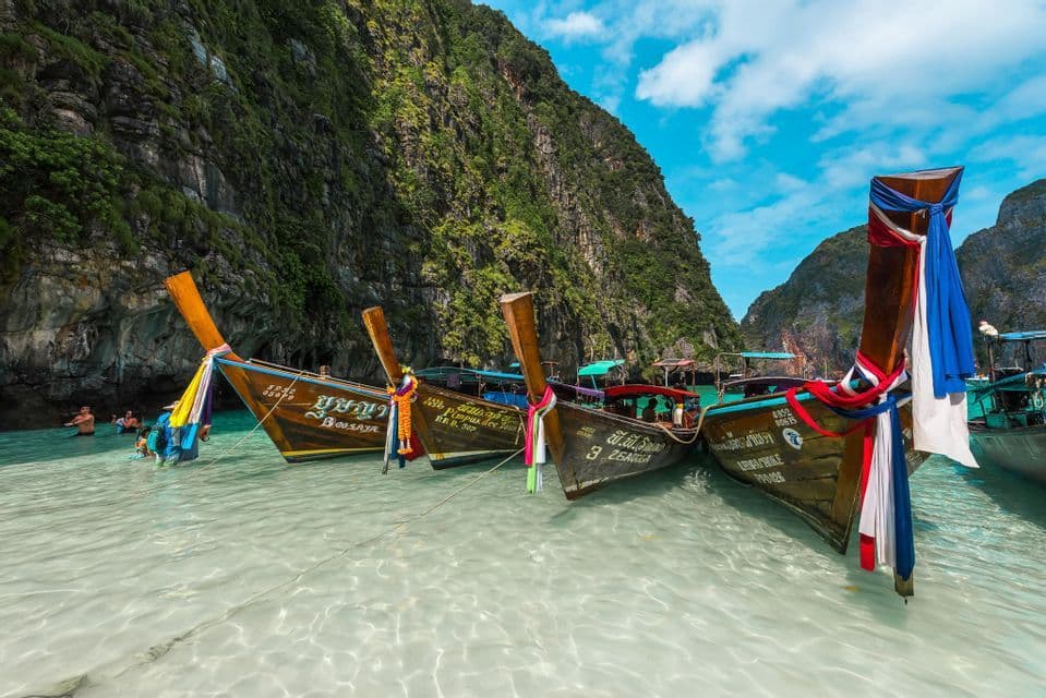 A WeRoad group trip swims in a clear lagoon next to traditional long-tail boats moored by steep green cliffs.