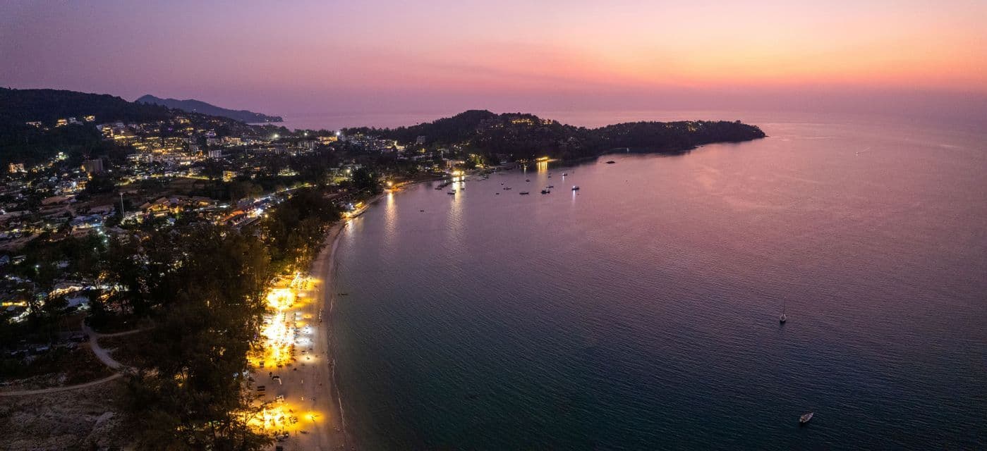 An aerial view of a brightly lit coastal town and beach next to a calm bay with boats at dusk.