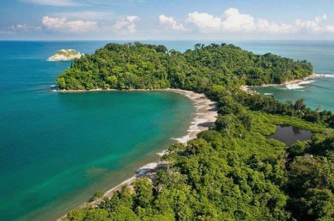 Vista aérea de una península tropical cubierta de densa vegetación, con una playa de arena curva y aguas oceánicas turquesas.
