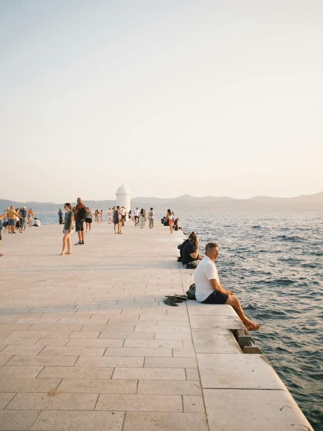 Ein Mann in einem weißen Hemd sitzt am Rande eines breiten Steinpiers und blickt auf das Wasser, während andere Menschen im Hintergrund spazieren gehen.