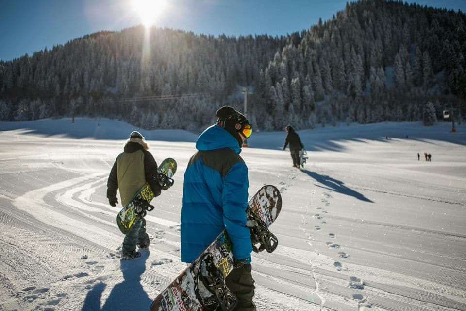 Un viaggio di gruppo WeRoad di persone che trasportano snowboard mentre salgono un pendio soleggiato e coperto di neve con una foresta sullo sfondo.