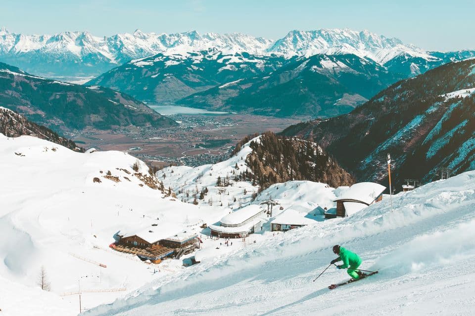 Uno sciatore in tuta verde scende un pendio innevato che domina una località di montagna, una valle e una lontana catena di cime innevate.