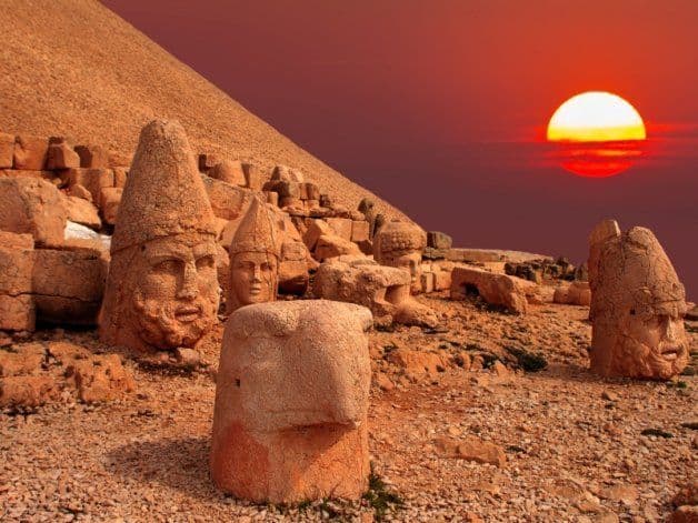 De grandes têtes de pierre antiques et des fragments de statues se dressent sur un flanc de montagne rocheux au coucher du soleil, sous un ciel rouge.