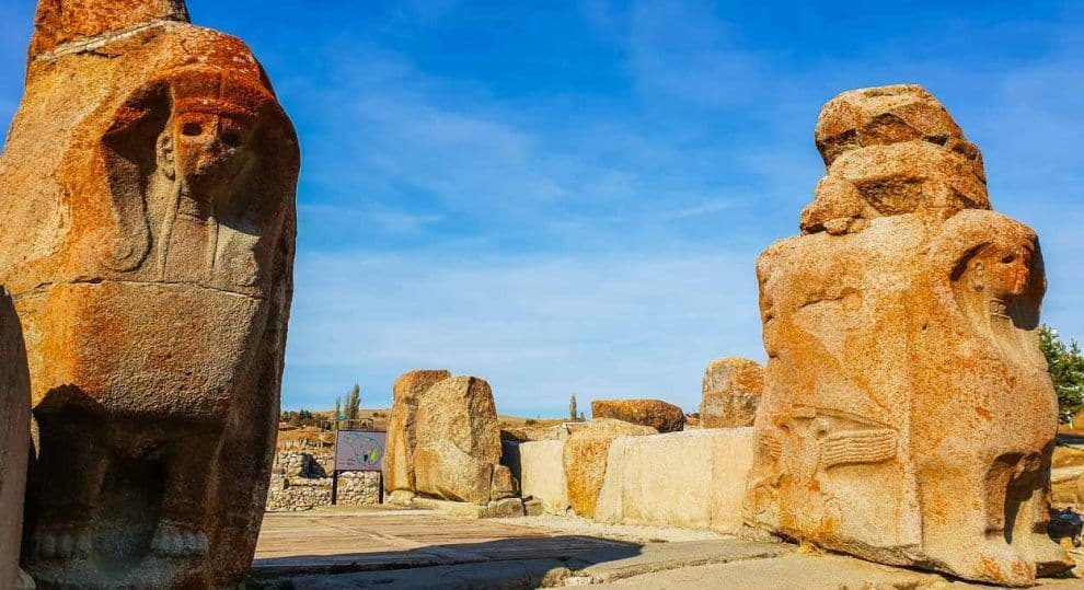 Deux grands monuments anciens en pierre rougeâtre avec des gravures se dressent sur un site archéologique sous un ciel bleu clair.