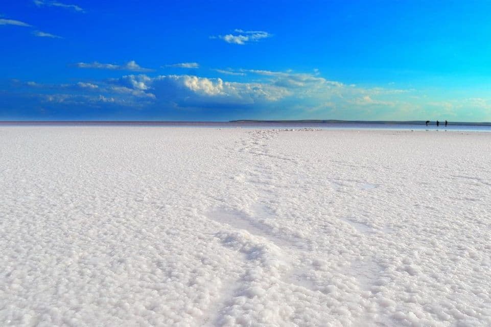 Des empreintes serpentent sur une étendue de sel blanc texturée vers trois silhouettes lointaines à l'horizon sous un ciel bleu éclatant.