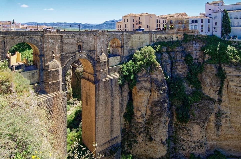 Un grande ponte ad arco in pietra attraversa un profondo canyon roccioso, collegando parti di una città storica costruita sulle scogliere.