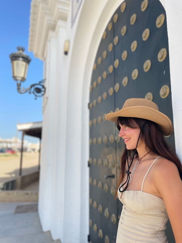 Una donna sorridente con un cappello di paglia si appoggia a un muro bianco accanto a una grande porta nera borchiata sotto un cielo blu.