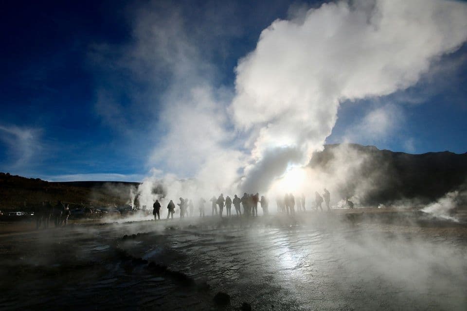 Sagome di un gruppo WeRoad osservano geyser eruttare vapore, illuminati dal sole splendente in un cielo blu.