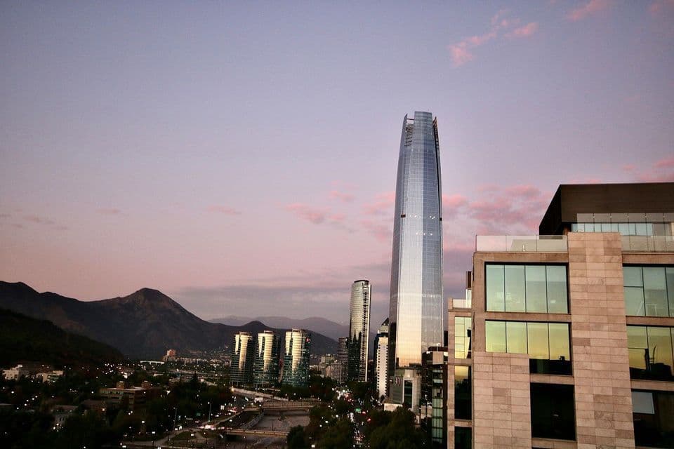 Uno skyline cittadino moderno con diversi grattacieli di vetro ai piedi di grandi montagne, durante un tramonto viola e rosa.