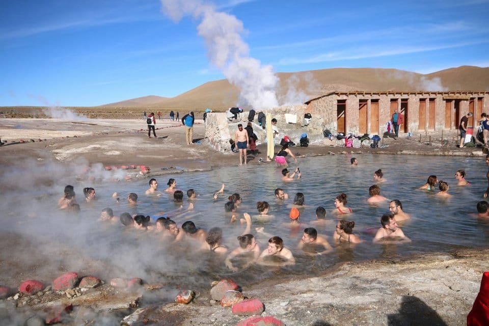 Un viaggio di gruppo WeRoad in relax in una sorgente termale all'aperto fumante con colline aride e un edificio in pietra sullo sfondo.