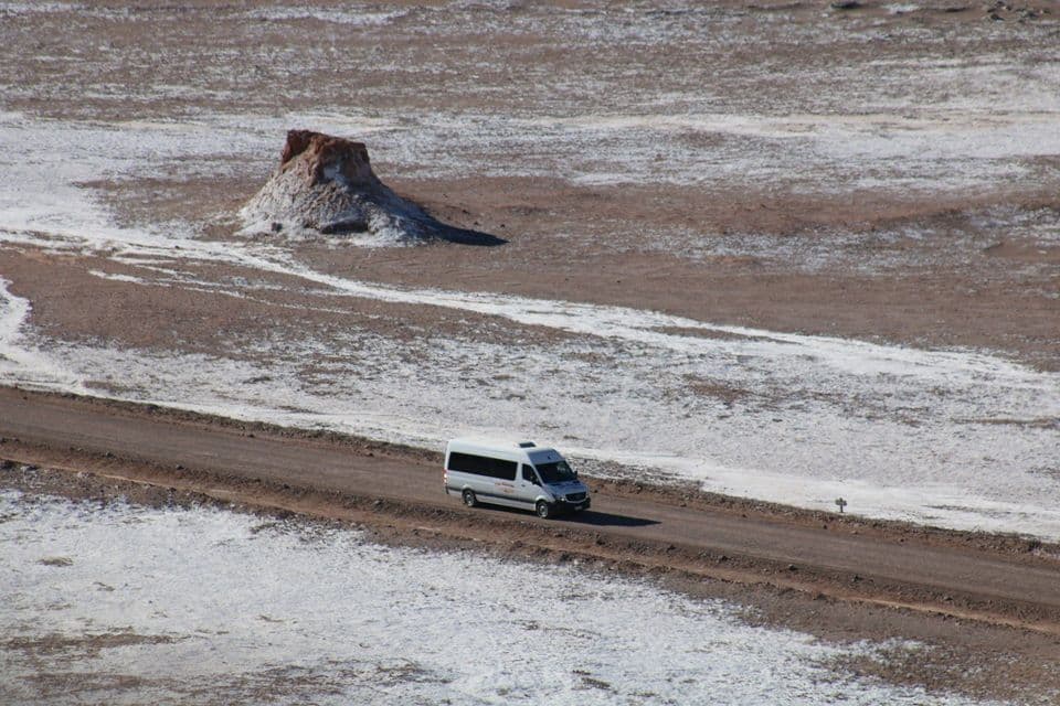 Un furgone bianco percorre una strada sterrata attraverso un vasto paesaggio coperto di sale con una formazione rocciosa solitaria.