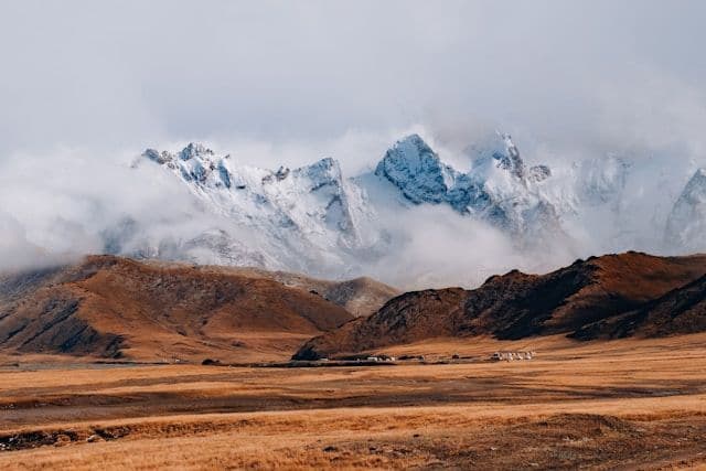 Picos de montañas nevadas se elevan entre las nubes sobre una vasta estepa dorada con un pequeño asentamiento en la distancia.