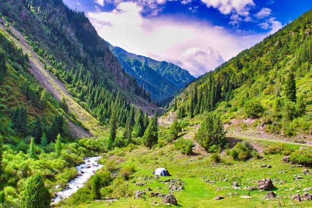 Una singola tenda è piantata su un prato verde accanto a un fiume in una vasta valle circondata da montagne ripide e alberate.