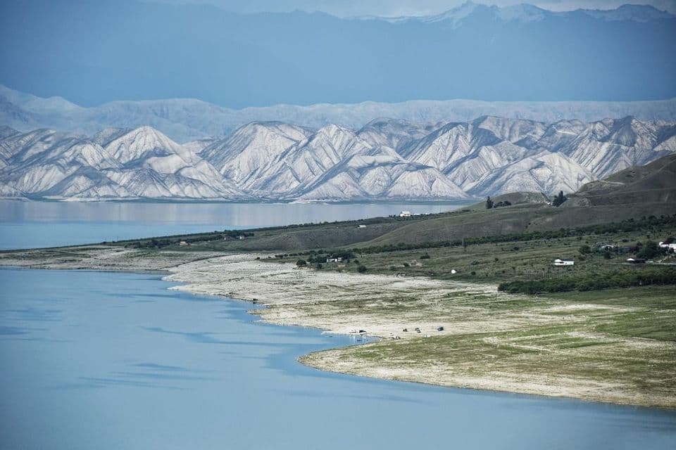 Un vasto lago blu si curva lungo una costa di verdi colline, con strati di catene montuose bianche e blu sullo sfondo.