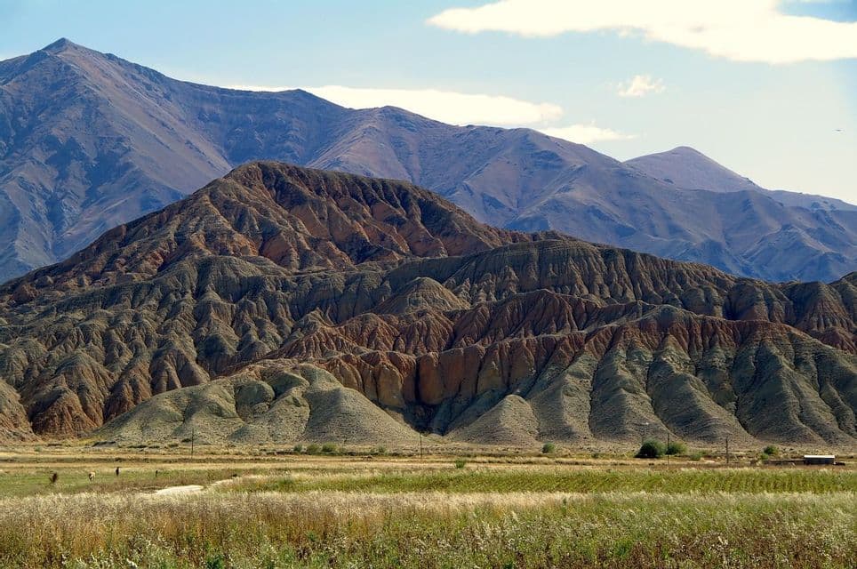 Colinas erosionadas con capas de roca roja y verde se alzan frente a una gran cordillera, con una llanura cubierta de hierba en primer plano.