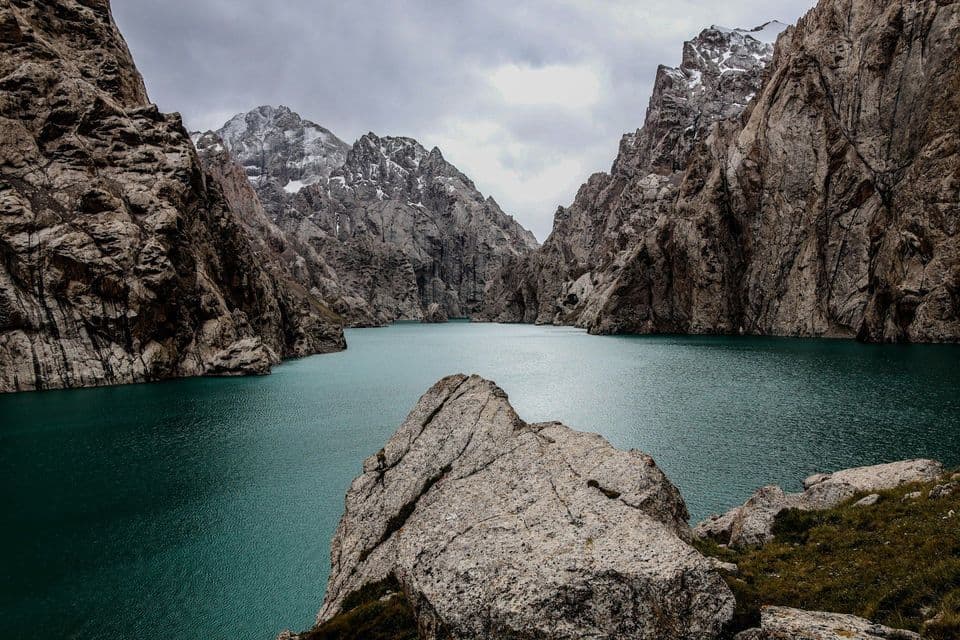 A turquoise lake fills a canyon surrounded by steep, rocky mountains with snow-capped peaks under a cloudy sky.