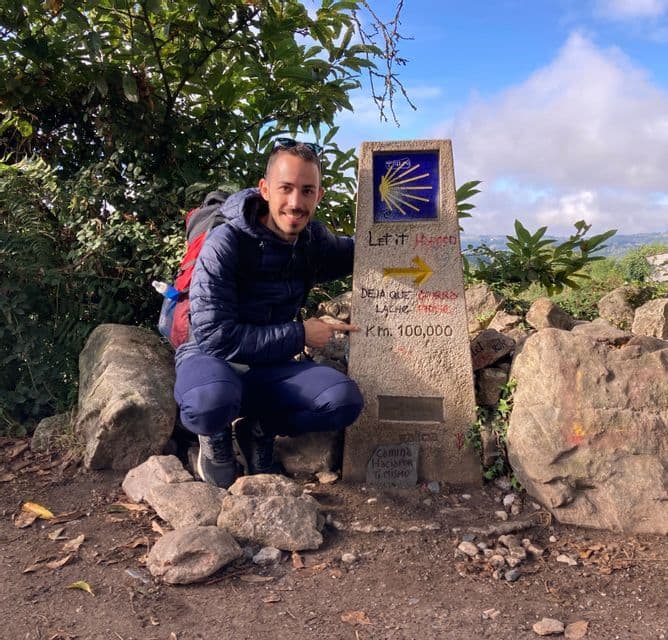 Ein lächelnder Wanderer mit Rucksack kniet auf einem Wanderweg und zeigt auf einen Steinmarker, auf dem 'Km. 100.000' steht.