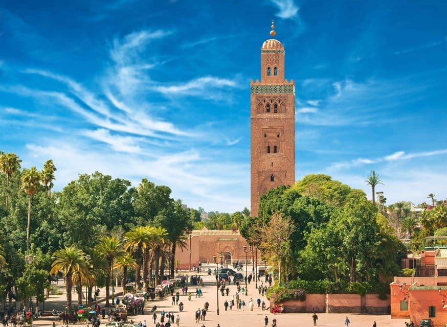 A tall, sand-colored minaret towers over a bustling city square filled with people and palm trees under a blue sky.