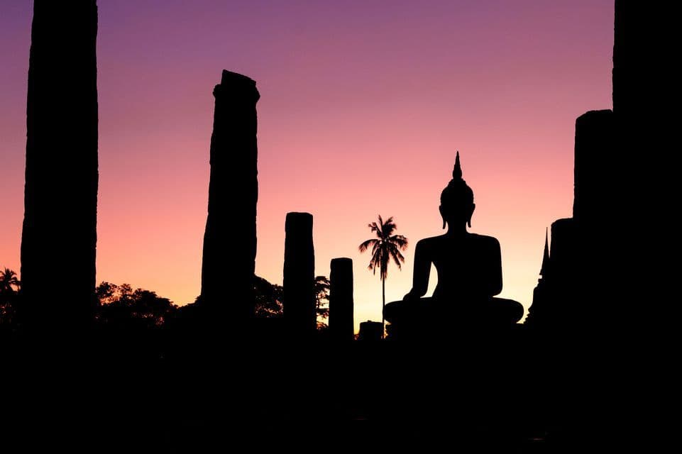 Eine Silhouette einer sitzenden Buddha-Statue zwischen antiken Ruinen und einer Palme vor einem lila-rosa Sonnenuntergangshimmel.