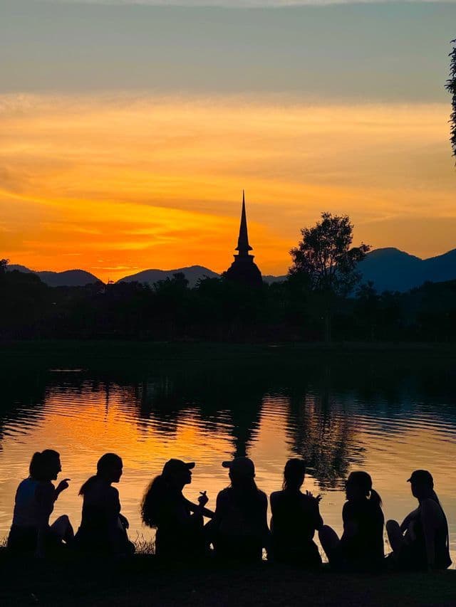 Un gruppo WeRoad in viaggio si staglia contro un lago, ammirando un tramonto arancione dietro un tempio lontano e le montagne.