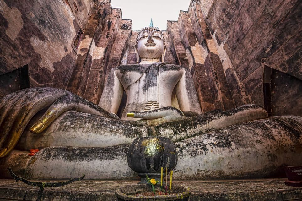 Eine kolossale, verwitterte Stein-Buddha-Statue sitzt meditierend zwischen hohen Steinmauern, mit einer kleinen Opferschale davor.