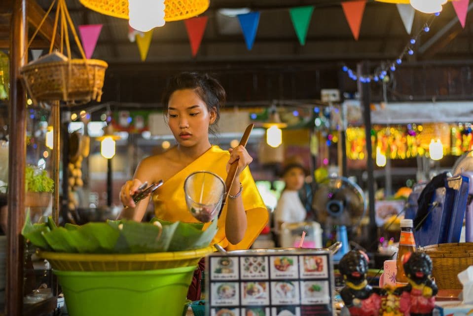 Una donna in top giallo prepara un pasto con uno scolapasta in una bancarella di cibo ben illuminata in un mercato coperto.