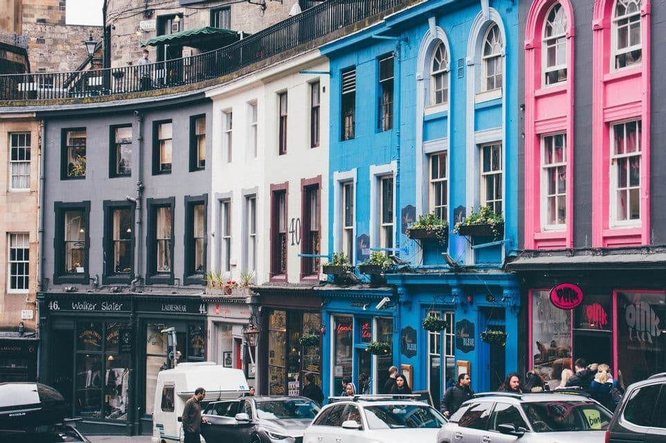 Eine Reihe bunter Gebäude mit Fassaden in Grau, Weiß, Blau und Rosa an einer geschwungenen Straße mit Geschäften, Fußgängern und geparkten Autos.
