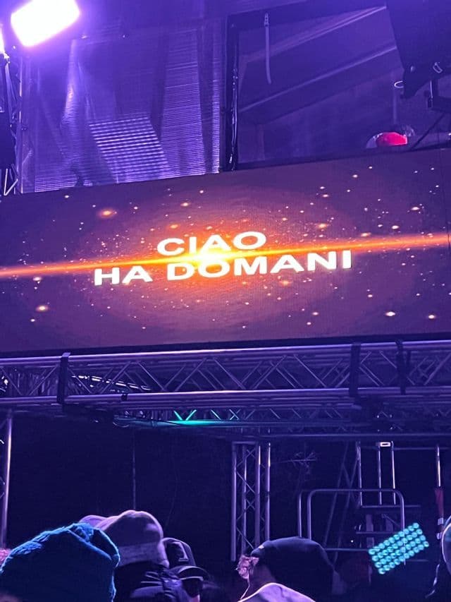 Un grande schermo durante un evento notturno mostra il testo 'CIAO HA DOMANI' su uno sfondo viola e cosmico, montato su una struttura metallica da palco.