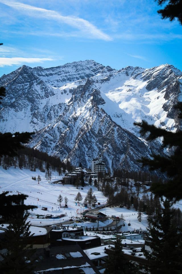 A high-angle view of a ski resort village in a snowy valley, with towering, snow-capped mountains in the background under a blue sky.