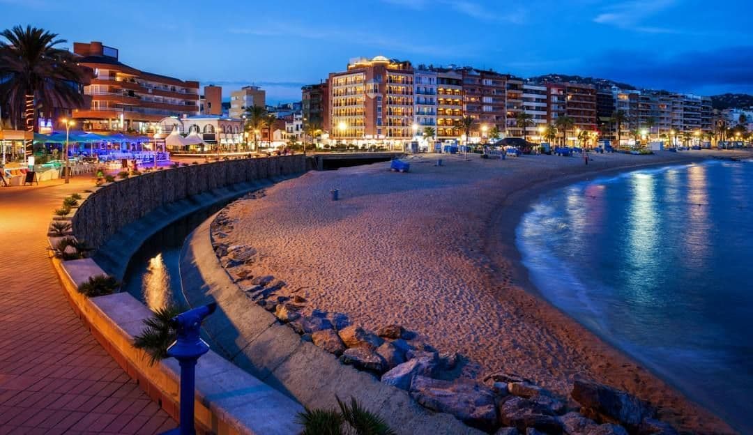 Una passeggiata cittadina illuminata si snoda lungo una spiaggia sabbiosa e il mare al crepuscolo, con le luci degli edifici che si riflettono sull'acqua.