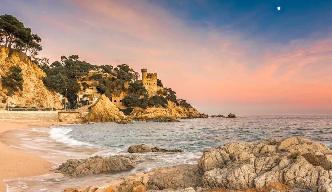 Un castello di pietra si trova su una scogliera rocciosa e alberata sopra una spiaggia sabbiosa e il mare sotto un cielo al tramonto rosa e blu.