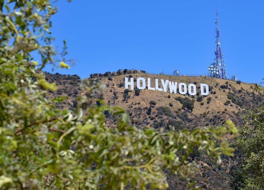 The Hollywood sign on a sunlit hill with a radio tower, viewed through out-of-focus green foliage against a clear blue sky.