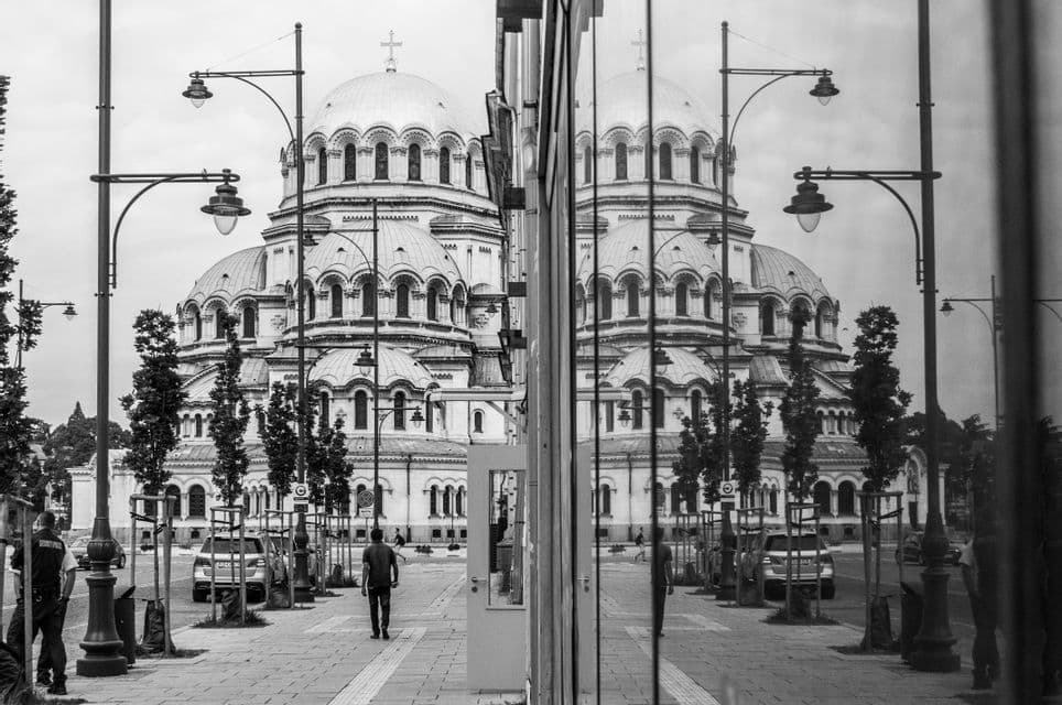 Una gran catedral con cúpula se refleja en la fachada de cristal de un edificio, creando una escena callejera simétrica en blanco y negro.