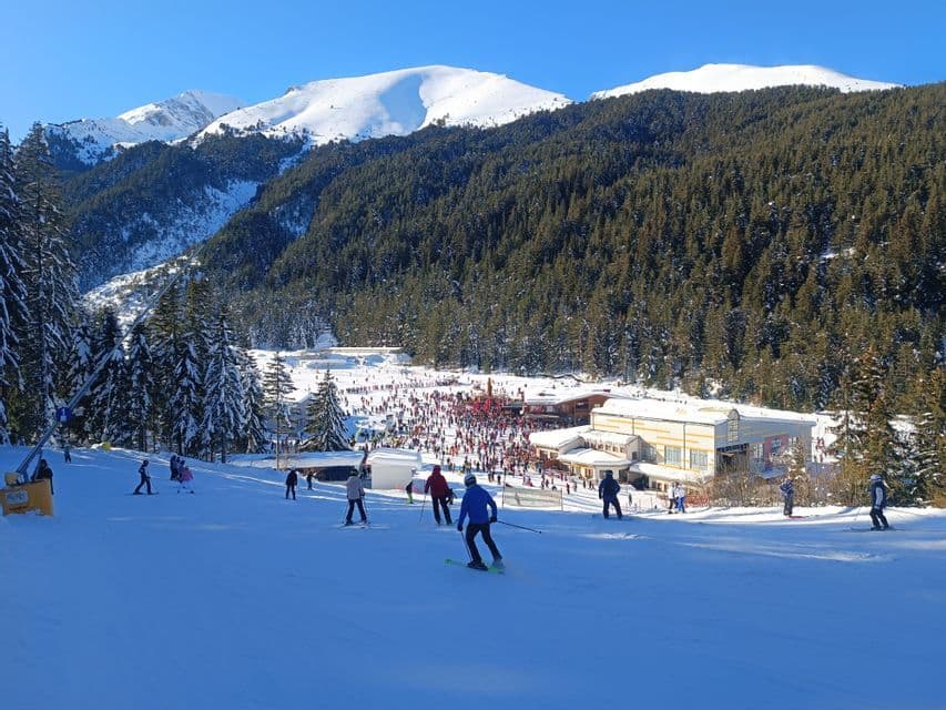 Una vista panorámica de una concurrida estación de esquí con gente en las pistas, un refugio al pie y montañas nevadas al fondo.