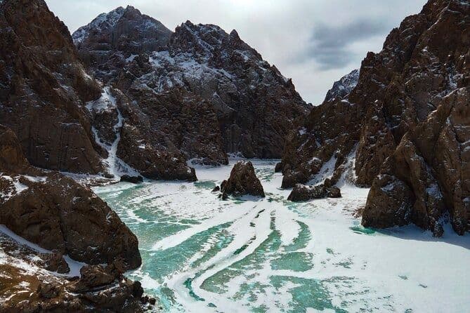 A rocky canyon with snow-dusted peaks surrounds a frozen river showing cracks of turquoise-colored ice.