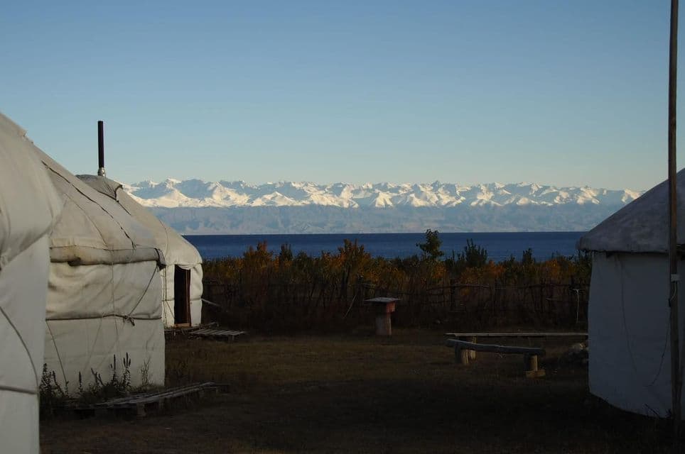 Yurtas blancas a la orilla de un lago con una cordillera nevada en la distancia bajo un cielo despejado.