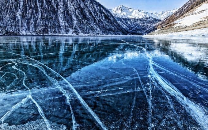 La glace bleue et claire d'un lac gelé est fissurée, reflétant une forêt et des montagnes enneigées.