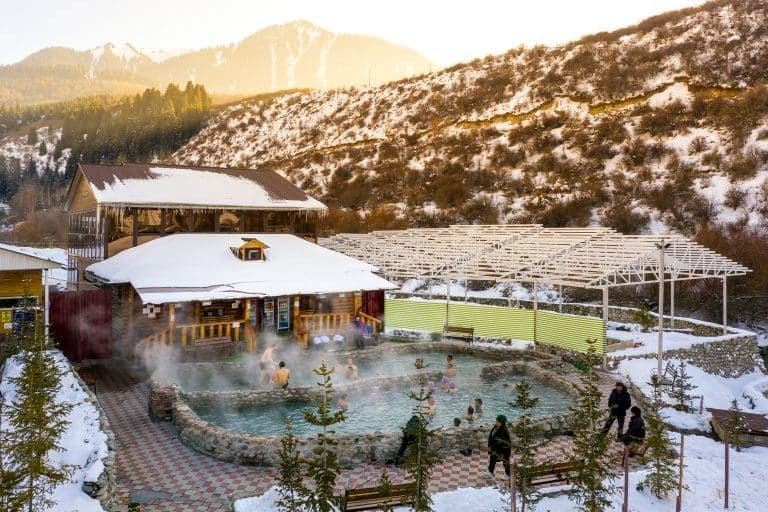 A WeRoad group trip relaxing in a steaming outdoor hot spring, surrounded by snow-covered cabins and mountains.