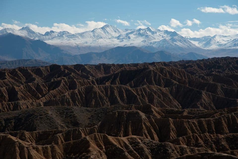 Eine Landschaft aus braunen, erodierten Hügeln im Vordergrund mit einer Kette schneebedeckter Berge in der Ferne unter blauem Himmel.