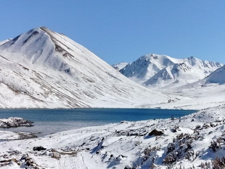 Un lago alpino blu si trova alla base di grandi montagne innevate sotto un cielo azzurro chiaro.