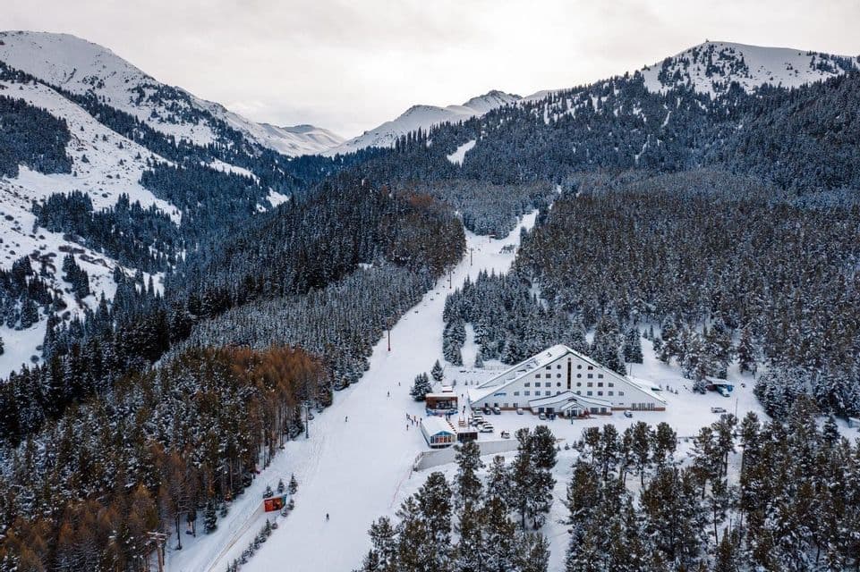 Eine Luftaufnahme eines Skigebiets mit einem großen Hotel am Fuße einer Piste, umgeben von schneebedeckten Bergen und Pinienwäldern.