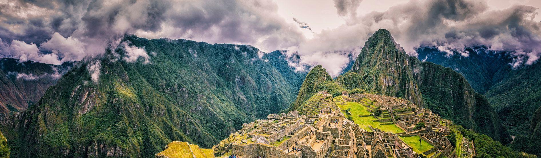 A panoramic view of ancient stone ruins on a mountain, surrounded by lush green peaks under a dramatic, cloudy sky.