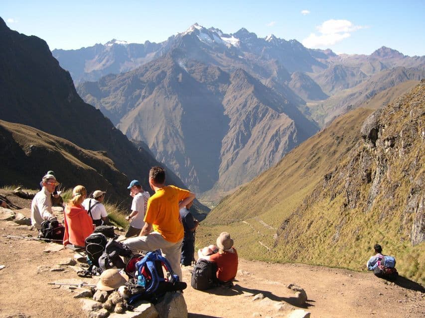 A WeRoad group trip of hikers rests on a mountain trail, looking out over a deep valley and distant snow-capped peaks.