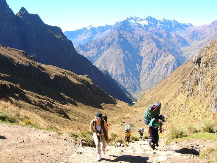 A WeRoad group trip hikes up a steep, dusty trail in a mountain valley with snow-capped peaks in the distance.