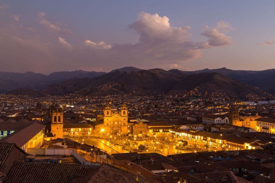 A historic city's illuminated central plaza and church seen from above at dusk, with mountains in the background.