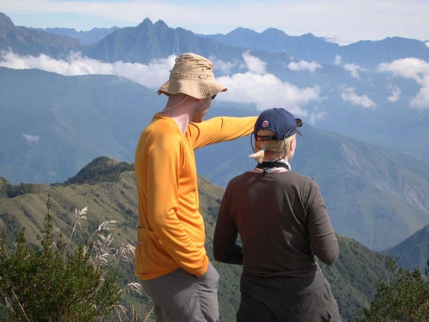 Two people from a WeRoad group trip stand on a mountain viewpoint, with one pointing at the distant peaks under a blue sky.