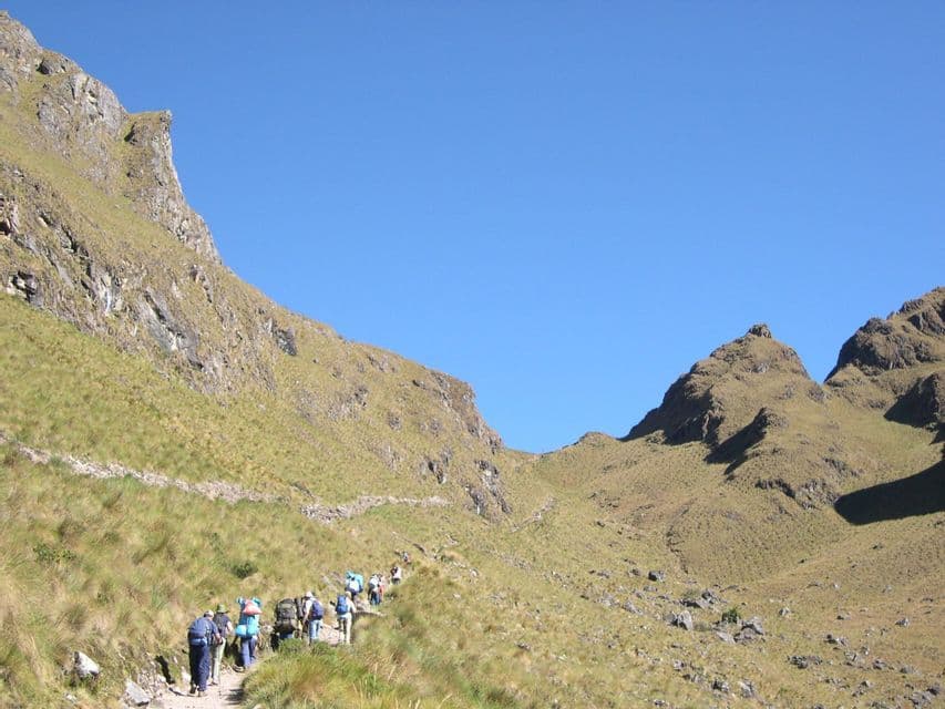 A WeRoad group trip hikes in a line on a narrow path up a steep, grassy mountain under a clear blue sky.