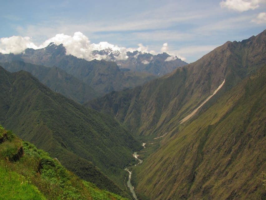 A river winds through a deep, green valley surrounded by mountains with snow-capped peaks in the distance.