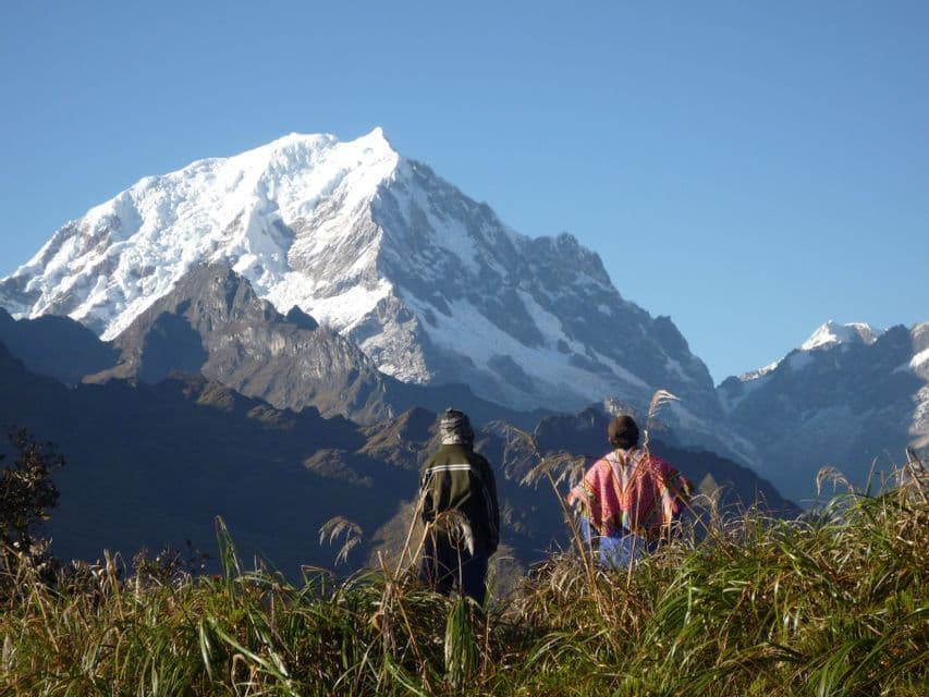 Two people from a WeRoad group trip stand in tall grass looking at a large, snow-capped mountain range against a clear blue sky.